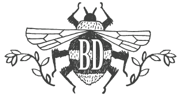 Brooklyn Doran logo image. A drawn bumblebee with laurels on either side. In the body of the bee is the initials "BD"